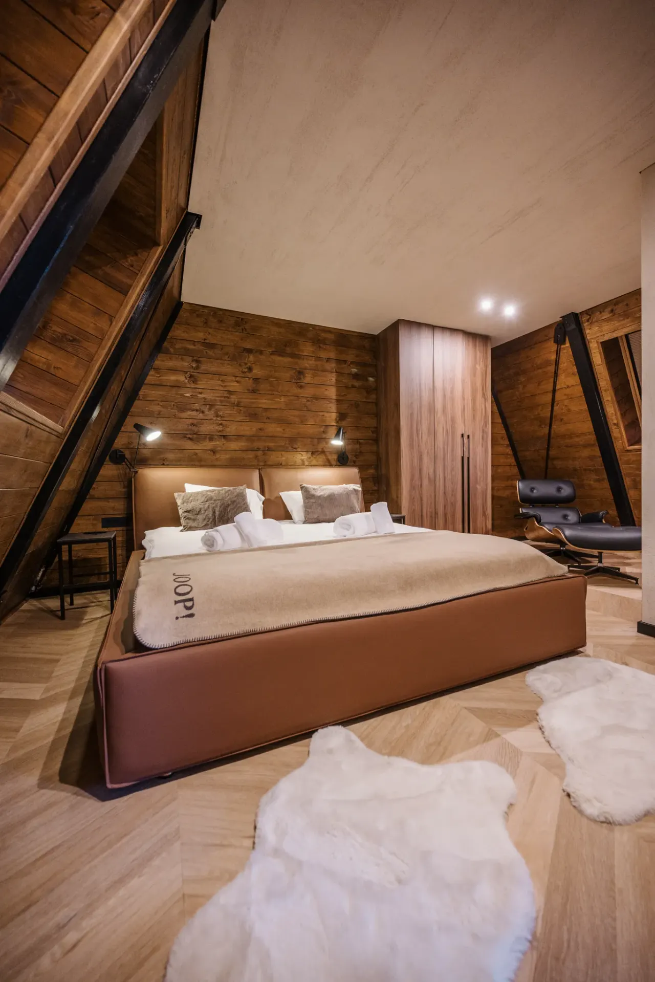 Fully wood-clad bedroom seen across its width, lounge chair and wardrobe beyond the bed